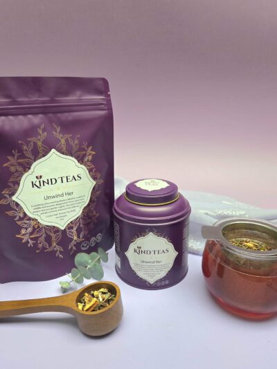 Purple Kind Teas packaging and canister with gold detailing, set beside a copper teapot and a wooden scoop of loose-leaf tea on a white surface.