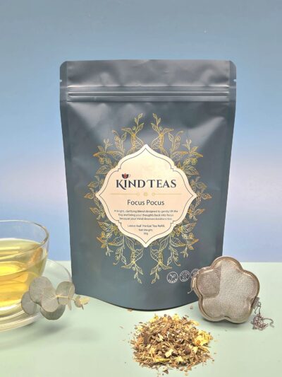 Kind Teas Focus Focus packaging with gold floral design, loose leaves scattered, a glass cup of tea, and a metal infuser nearby