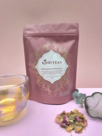 Pink resealable tea pouch labeled Kind Teas 'Menopause Moments' with a glass cup of tea and loose herbal tea leaves nearby on a pale pink background.