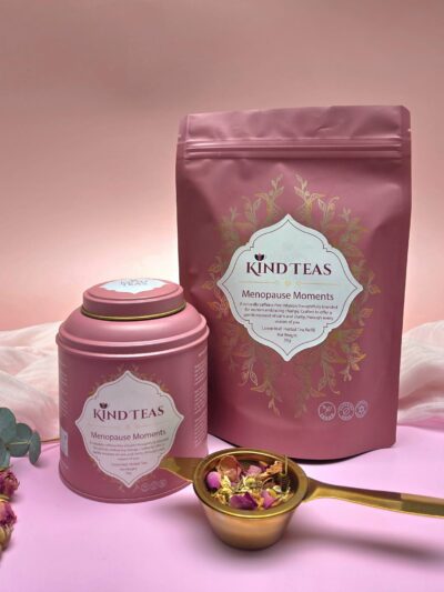 Pink tea packaging set labeled 'Kind Teas Menopause Moments' with a matching round tin, standing pouch, and a gold scoop filled with dried petals on a pink backdrop.