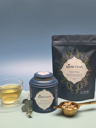 Kind Teas Focus Focus tea package and matching tin, with a glass teacup of brewed tea and a gold spoon of loose leaves on a white surface.