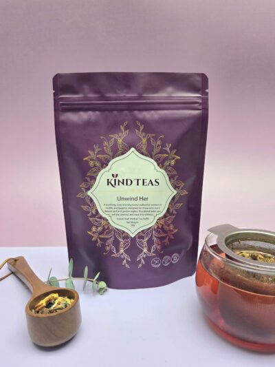 Purple Kind Teas pouch labeled 'Unwind Her' with an ornate herbal design; in front, a wooden scoop of loose tea and a metal infuser cup.