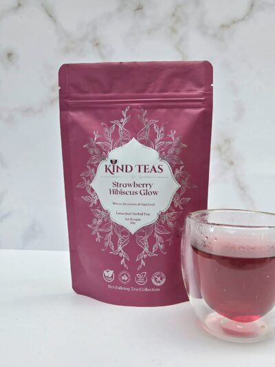 Pink Kind Teas pouch labeled Strawberry Hibiscus Glow with a glass of pink tea beside it on a light surface.
