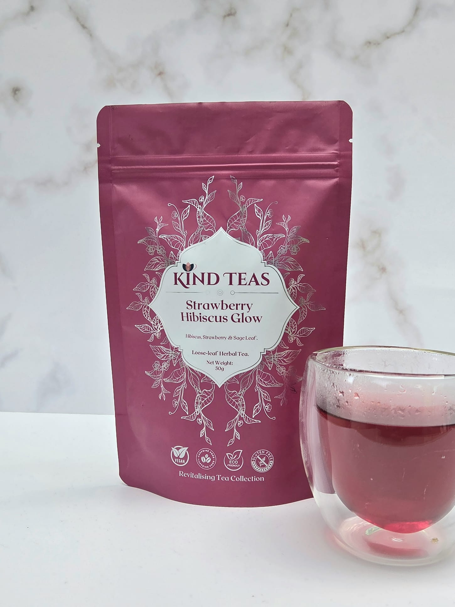 WhatsApp Image 2026-05-05 at 09.14.20 Strawberry Hibiscus Pink Kind Teas pouch labeled Strawberry Hibiscus Glow with a glass of pink tea beside it on a light surface.