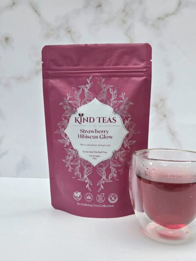 Pink tea package labeled Kind Teas Strawberry Hibiscus Glow with a white decorative label and floral design, standing beside a glass of pink tea.