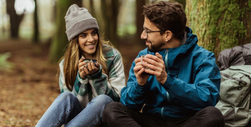 Cheerful millennial caucasian woman and guy in jacket with backpack travel in cold forest, drink hot tea, enjoy adventure together outdoor. Walking, vacation, active lifestyle and camping, sports